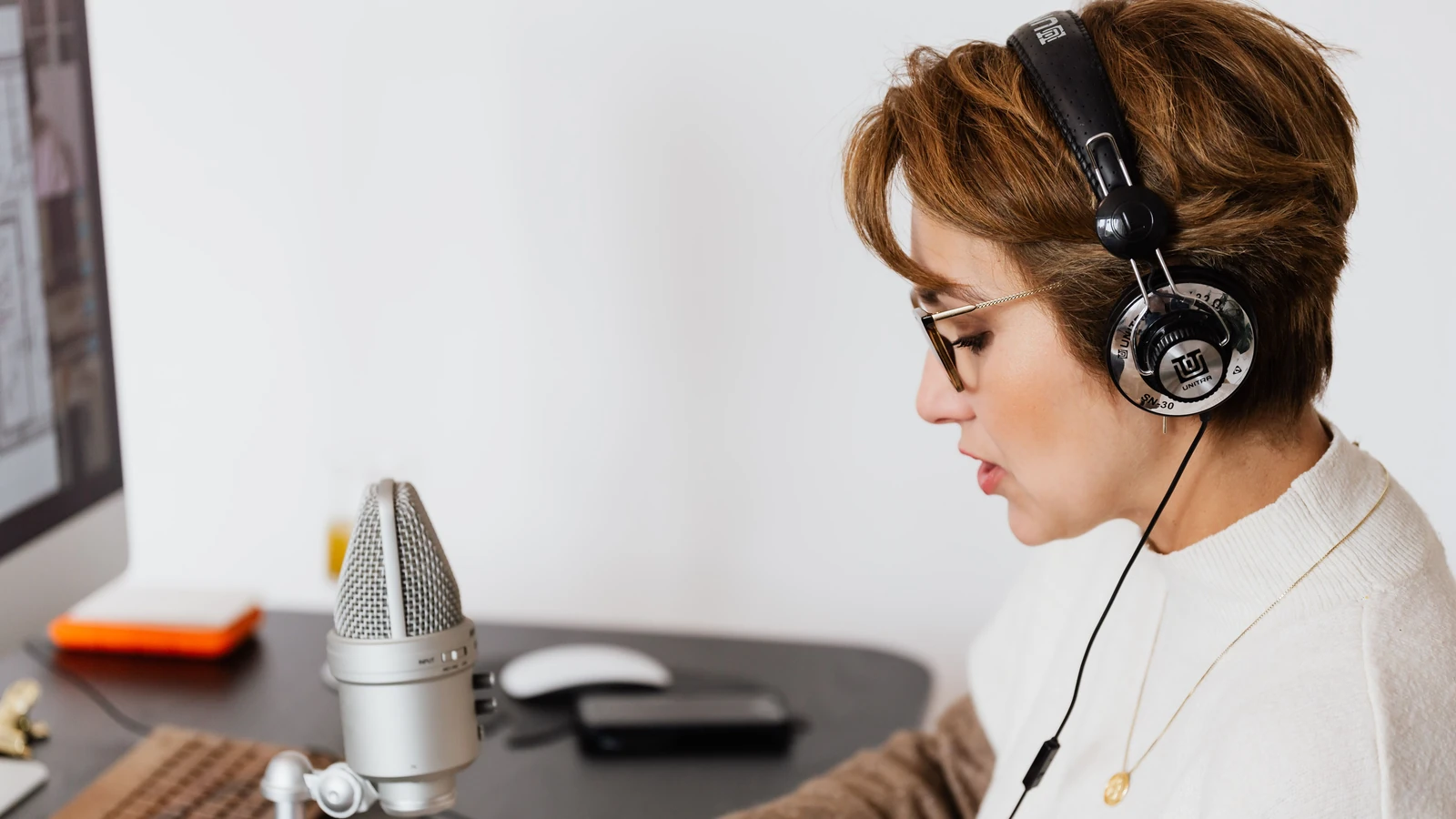 Una mujer con auriculares y gafas trabaja ante un micrófono en un estudio doméstico.