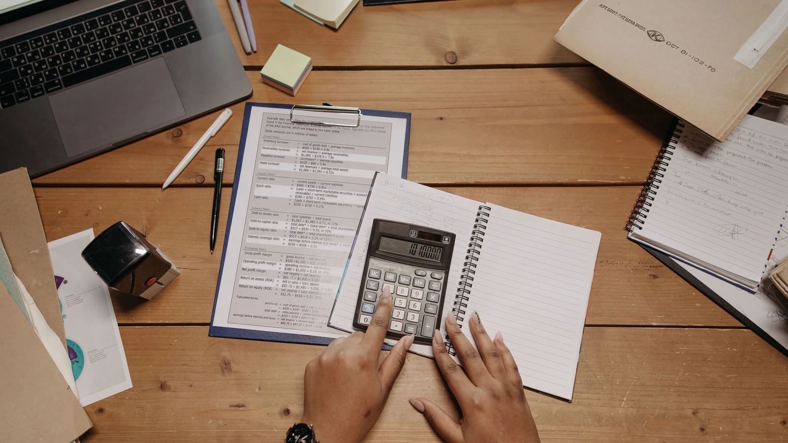Unas manos utilizan una calculadora junto a documentos impresos y un portátil sobre una mesa.