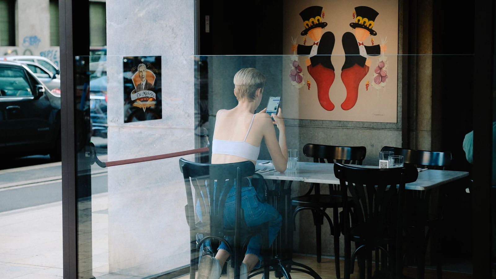 Una mujer consulta su teléfono sentada en una cafetería, vista a través del ventanal.