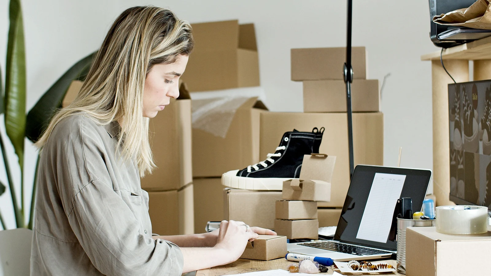 Una mujer prepara una caja junto a un portátil, paquetes y productos.