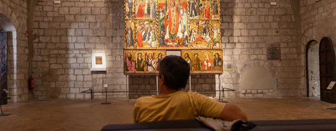 Una persona sentada contempla un retablo en una sala de piedra del Museu d’Art de Girona.