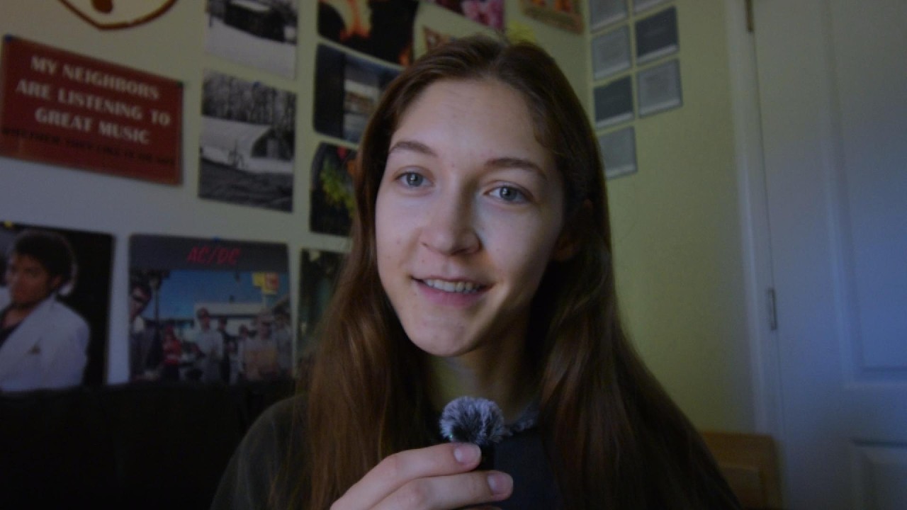 Emilymarie habla a cámara con un pequeño micrófono, ante una pared con carteles.
