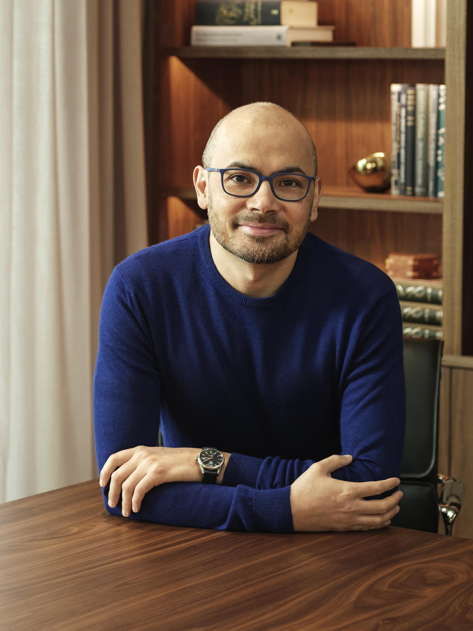 Demis Hassabis sentado ante una estantería, con jersey azul y gafas.