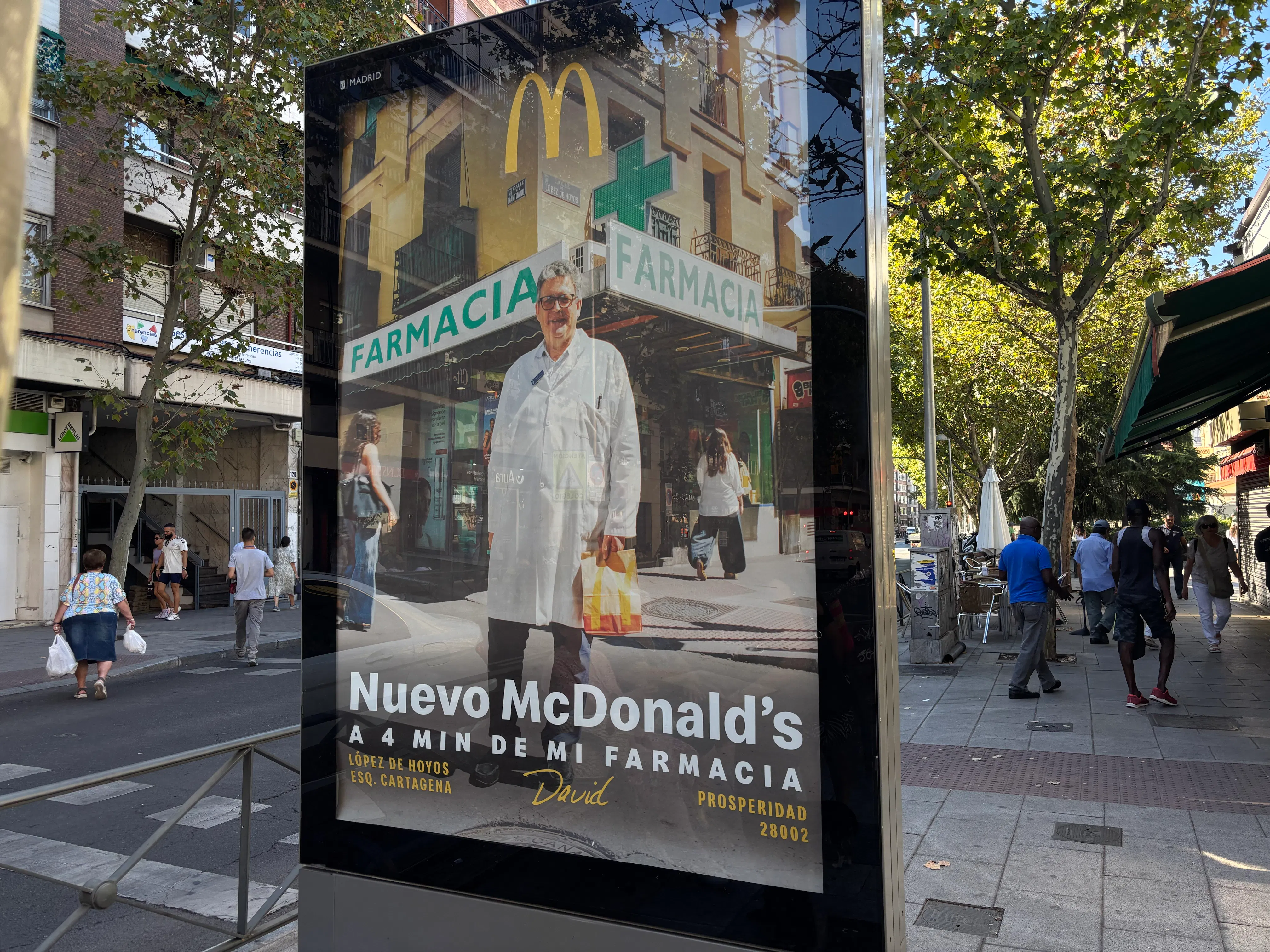 Mupi de la campaña de McDonald’s dedicado a una farmacia, instalado en una calle arbolada.